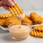 A close-up shot of a hand holding a waffle fry being dipped into a bowl of creamy Chick-fil-A Sauce . Waffle fries and chicken nuggets are visible on a plate in the background.