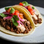 Two delicious tacos filled with seasoned ground beef are presented on a speckled plate. The tacos are topped with a creamy sauce, pickled red onions, chopped tomatoes, and fresh cilantro, suggesting a potential side or creative way to enjoy leftover, flavorful beef when thinking about how to cook wagyu ribeye. Although not a ribeye, the rich filling in the tacos implies the care taken in cooking high-quality wagyu beef. The presentation highlights how versatile and delicious wagyu can be, beyond traditional steak preparations.