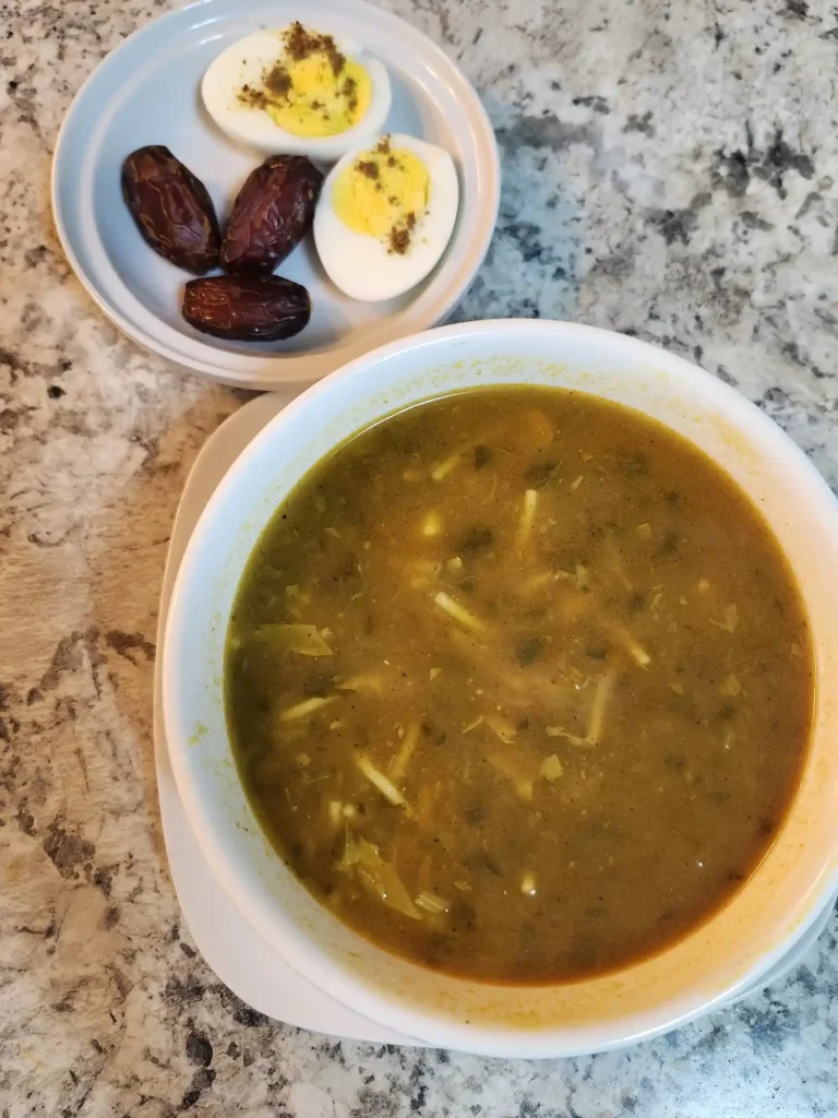 A bowl of Moroccan Harira soup served with noodles, chickpeas, and herbs, accompanied by a side plate with  boiled eggs, and dates.