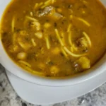 A bowl of Harira soup, a traditional Moroccan dish, featuring a rich tomato-based broth with chickpeas, lentils, noodles, and tender pieces of meat, garnished with herbs.