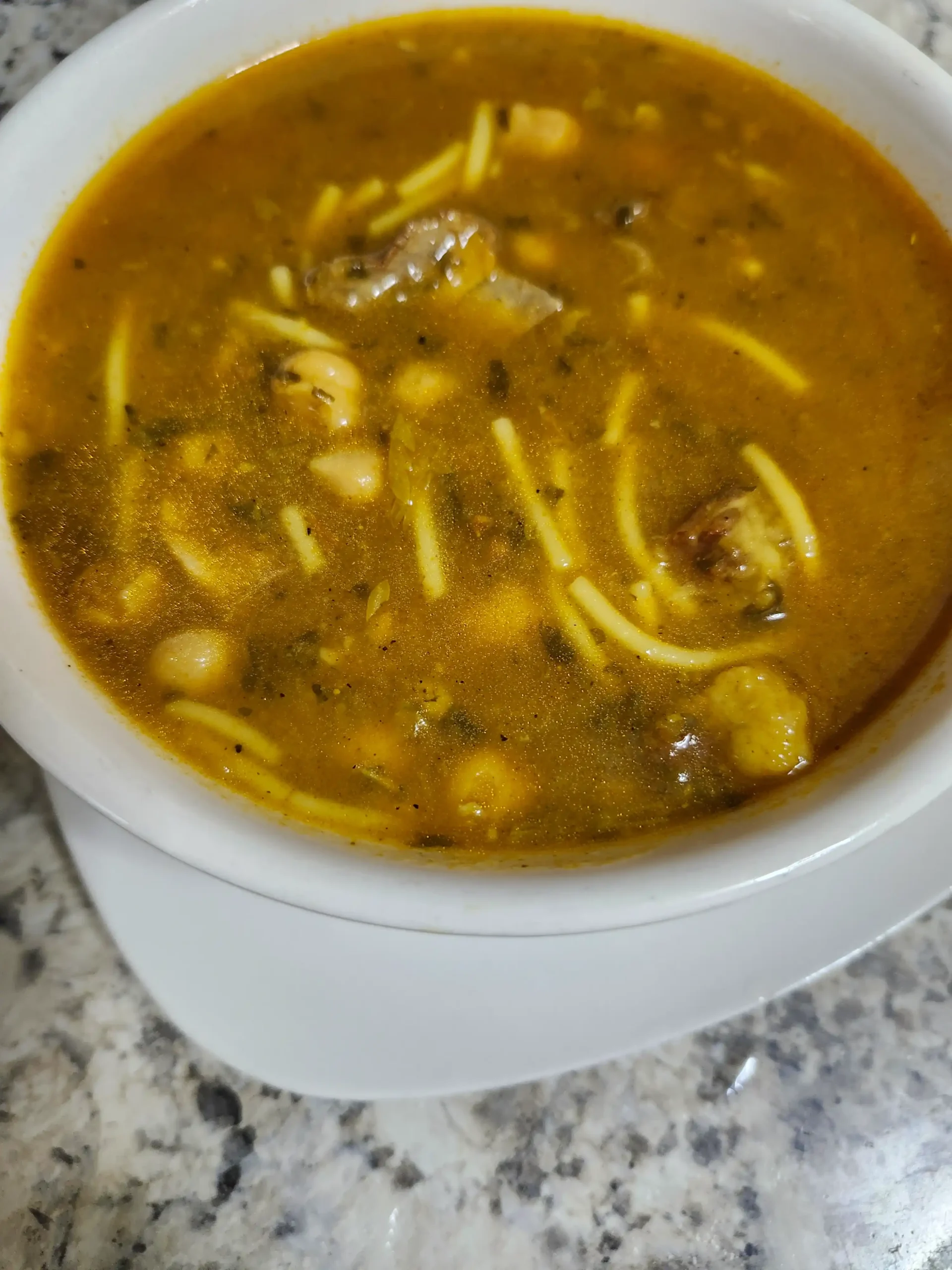 A bowl of Harira soup, a traditional Moroccan dish, featuring a rich tomato-based broth with chickpeas, lentils, noodles, and tender pieces of meat, garnished with herbs.