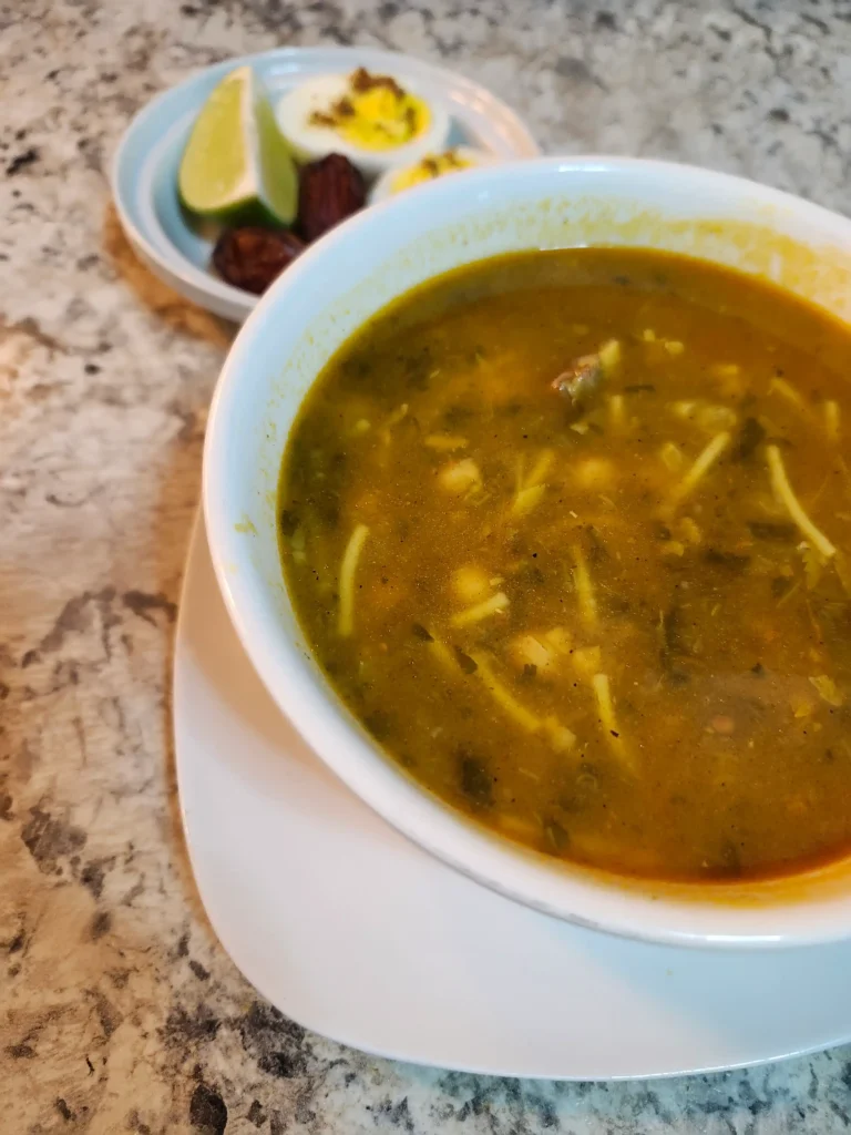 A bowl of Moroccan Harira soup served with noodles, chickpeas, and herbs, accompanied by a side plate with lime wedges, boiled eggs, and dates.