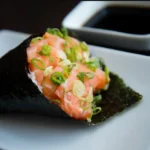 Close-up of a salmon temaki sushi cone with green onions, resting on a white plate; soy sauce dish in background