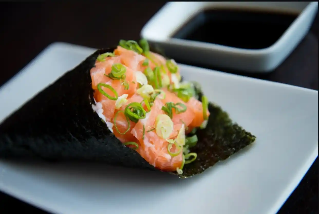 Close-up of a salmon temaki sushi cone with green onions, resting on a white plate; soy sauce dish in background