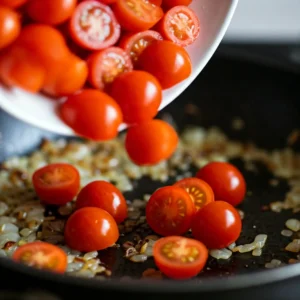 A close-up, slightly elevated shot of halved cherry tomatoes being added to a skillet already containing sautéed onions and garlic.