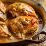 A close-up shot of Smothered Chicken Recipe in a rich, glossy gravy, served in a cast iron skillet. The chicken has a golden-brown crust.