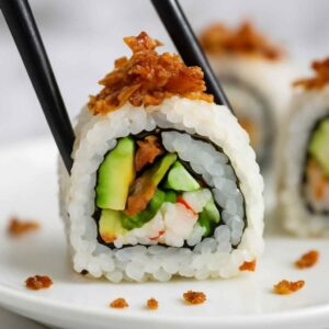 Close-up of a sushi roll held with black chopsticks, topped with crispy salmon skin, and filled with avocado, cucumber, and crab meat.