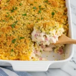 This image shows a close-up of a Chicken Cordon Bleu Casserole in a white baking dish. The casserole has a golden, crispy breadcrumb topping with finely chopped parsley sprinkled on top. A wooden spoon is lifting a serving, revealing creamy pasta mixed with diced smoked turkey, chicken, and possibly peas, coated in a cheesy, creamy sauce. The dish is set on a marble countertop with a blue and white striped kitchen towel partially visible.