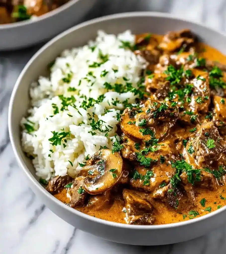 Bowl of creamy beef stroganoff with mushrooms and white rice, garnished with fresh parsley.