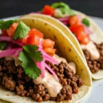 delicious wagyu ground beef tacos are arranged on a plate, garnished with a creamy sauce, pickled red onions, diced tomatoes, fresh cilantro, and thin radish slices. Lime wedges are visible on the side. The image showcases an appetizing dish of wagyu ground beef tacos, highlighting the vibrant colors and fresh ingredients used for a flavorful and satisfying meal.