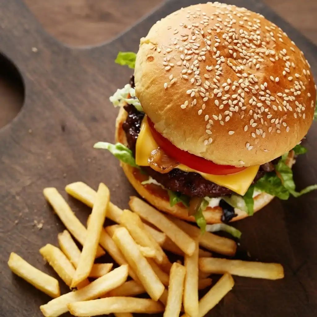 Gourmet Kobe beef burger with melted cheddar cheese, caramelized onions, lettuce, tomato, and a sesame seed bun, served with golden fries on a wooden board.