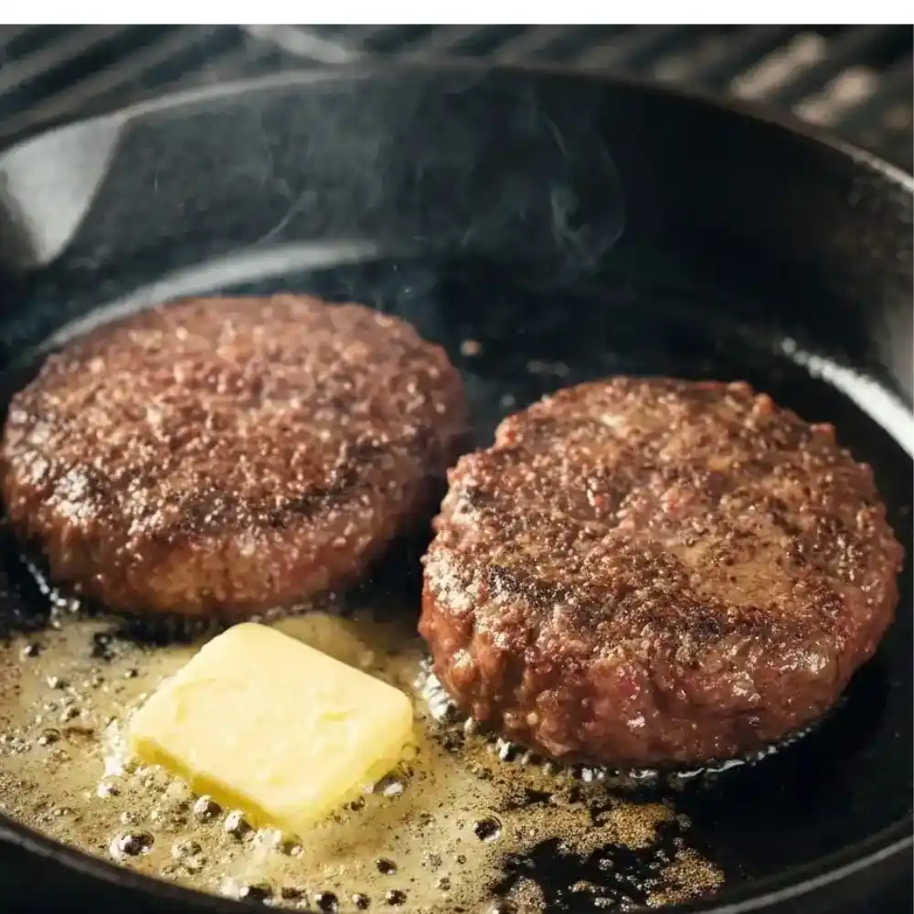 Kobe beef burger patties searing in a cast iron skillet with butter melting, on a grill with flames underneath.