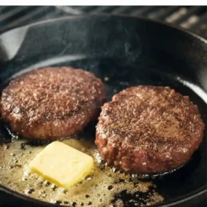 Kobe beef burger patties searing in a cast iron skillet with butter melting, on a grill with flames underneath.