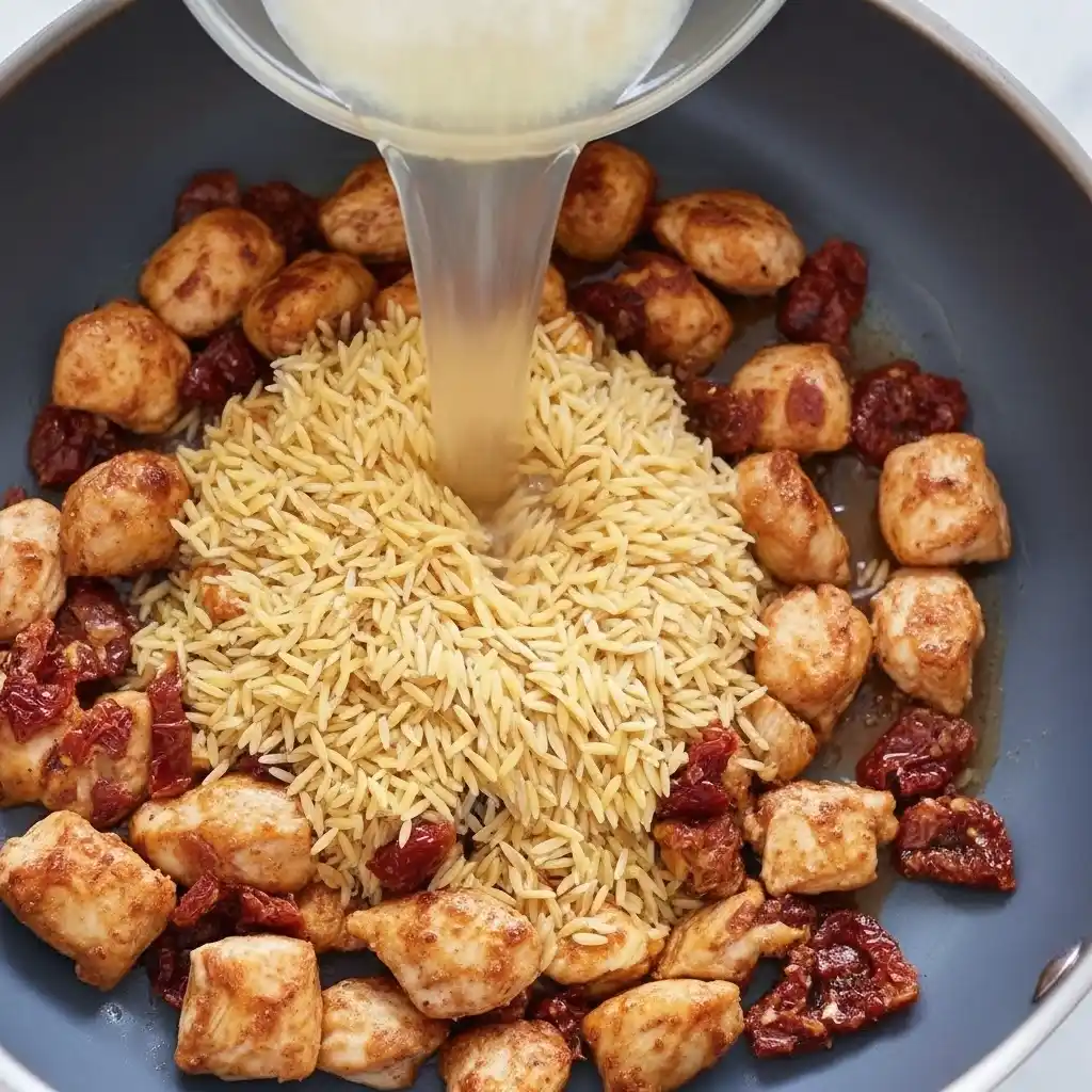 Orzo pasta and chicken with sun-dried tomatoes in a skillet with broth being poured in