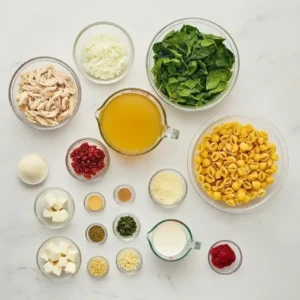 Fresh ingredients for Marry Me Chicken Soup including pasta shells, spinach, shredded chicken, broth, sun-dried tomatoes, onion, garlic, cream, and Parmesan laid out on a white surface.