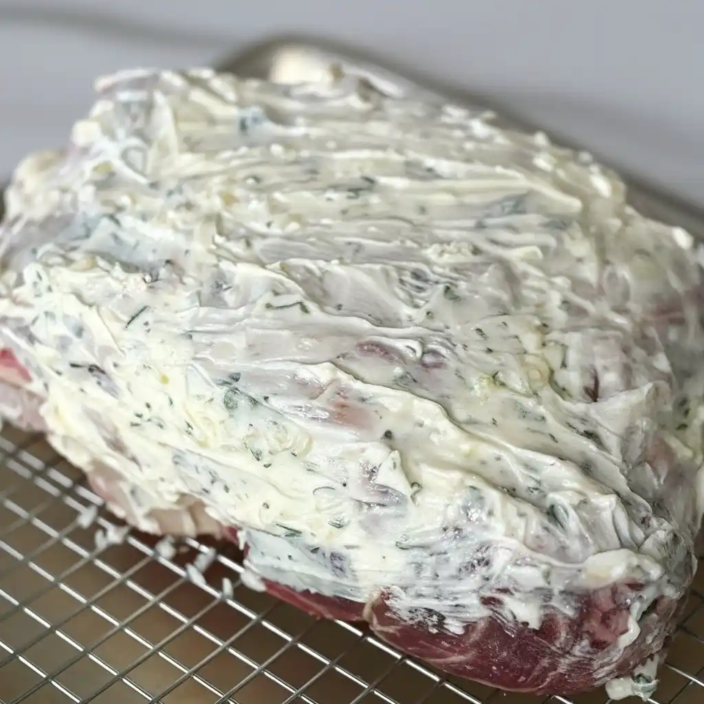 Prime rib roast fully coated in garlic-herb butter, resting on a wire rack over a baking tray.