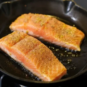 Texas Roadhouse-style salmon searing in a cast iron skillet with olive oil and spices.
