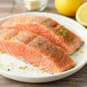 Raw salmon fillets seasoned with lemon zest, salt, and black pepper on a white plate.
