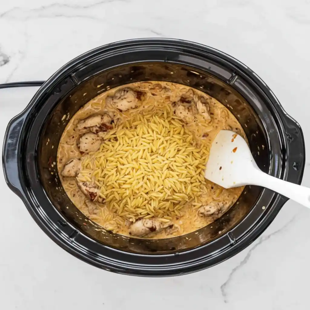 Adding orzo pasta to creamy chicken mixture in crockpot