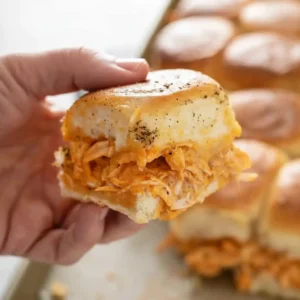 Hand holding a Buffalo Chicken Slider filled with shredded chicken, buffalo sauce, and melted cheese on a buttery roll.
