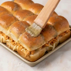 Brushing melted butter and Italian seasoning over Hawaiian rolls filled with buffalo chicken and cheese.